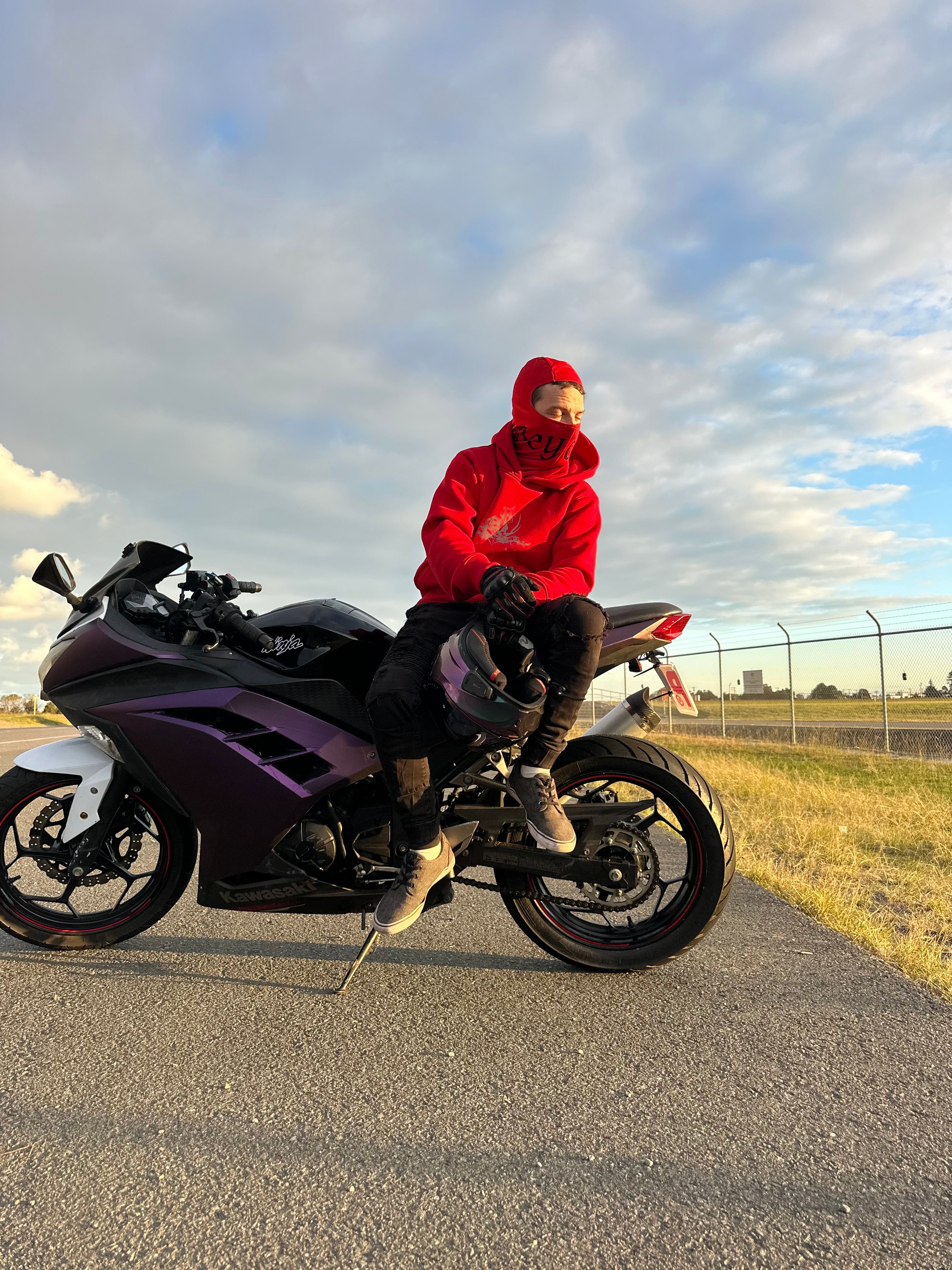 A person in a red hoodie with an inbuilt balaclava and black pants sits on a purple and black sport motorcycle parked on an asphalt road under a cloudy blue sky.