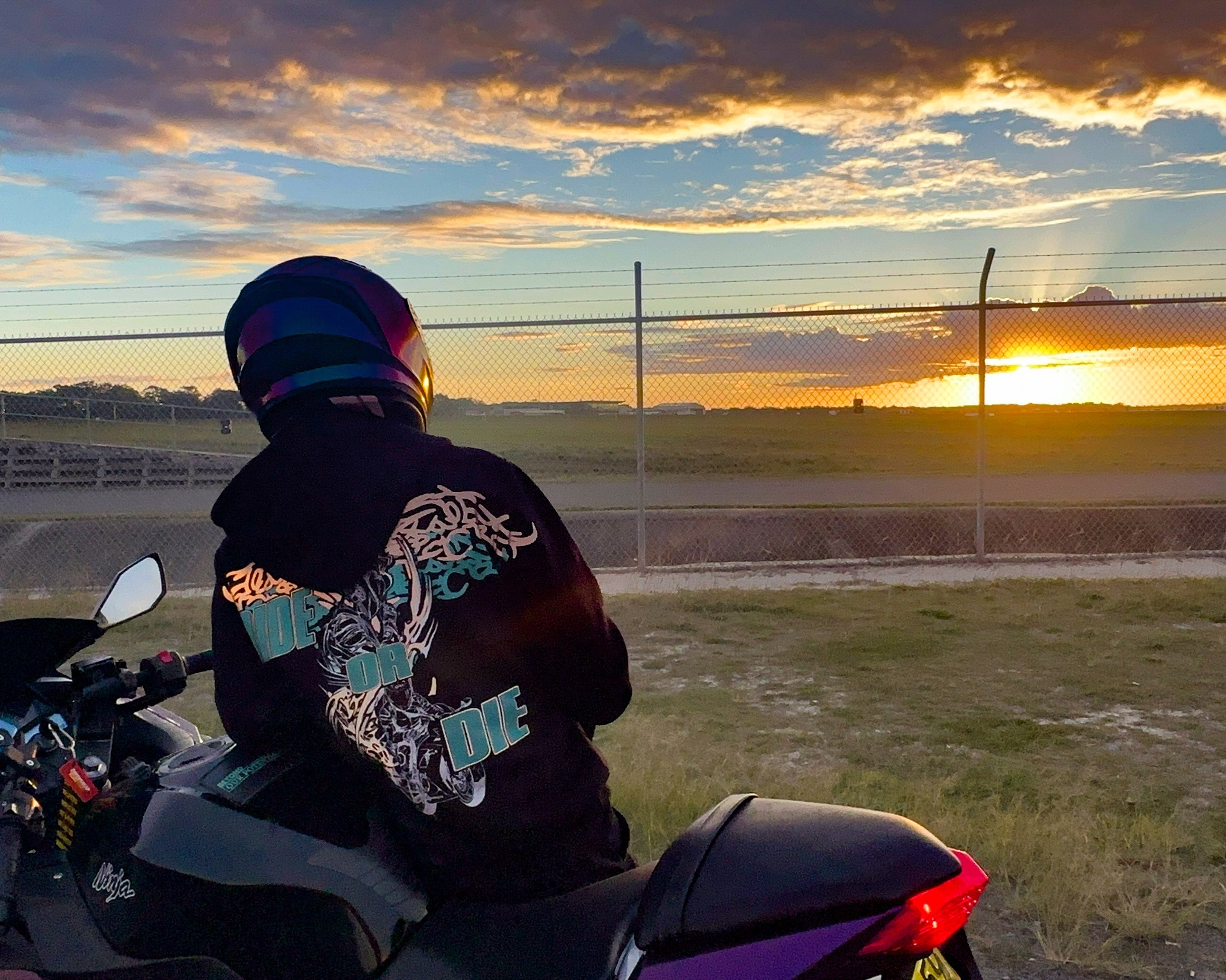 A person wearing a black graphic reflective hoodie and a colourful helmet sits on a purple and black motorcycle, facing a sunset over an open field with a fence in the background.