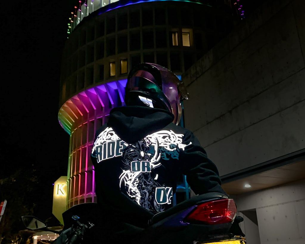 A motorbike rider wearing a black hoodie with a large reflective graphic print on the back sits on a motorcycle at night in front of a brightly lit cylindrical building with rainbow-colored lights.