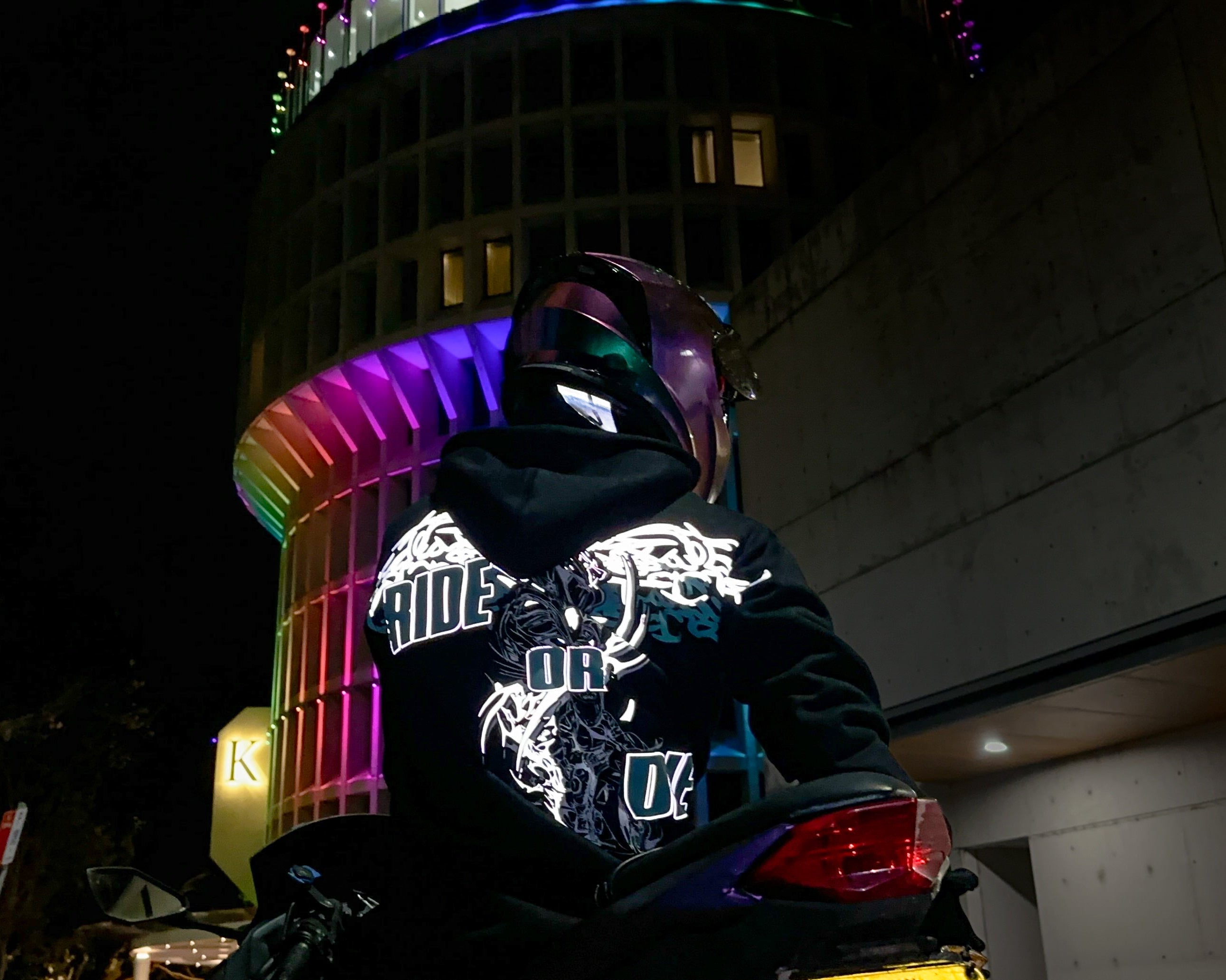 A motorbike rider wearing a black hoodie with a large reflective graphic print on the back sits on a motorcycle at night in front of a brightly lit cylindrical building with rainbow-colored lights.
