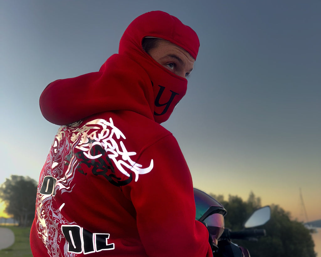 A rider wearing a bright red hooded jumper with an inbuilt balaclava sits on a motorcycle outdoors at dusk. The back of the jacket features large reflective panelling and black graphic text and dragon-style artwork