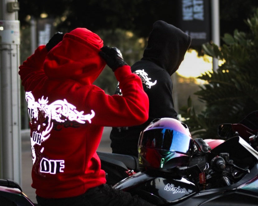 A person wearing a bright red hoodie with a large reflective design graphic on the back sits on a motorcycle, while another person in a black hoodie stands beside them. A motorcycle with a blue and pink helmet rests on the bike, and an outdoor street scene with trees and a hanging banner is visible in the background.
