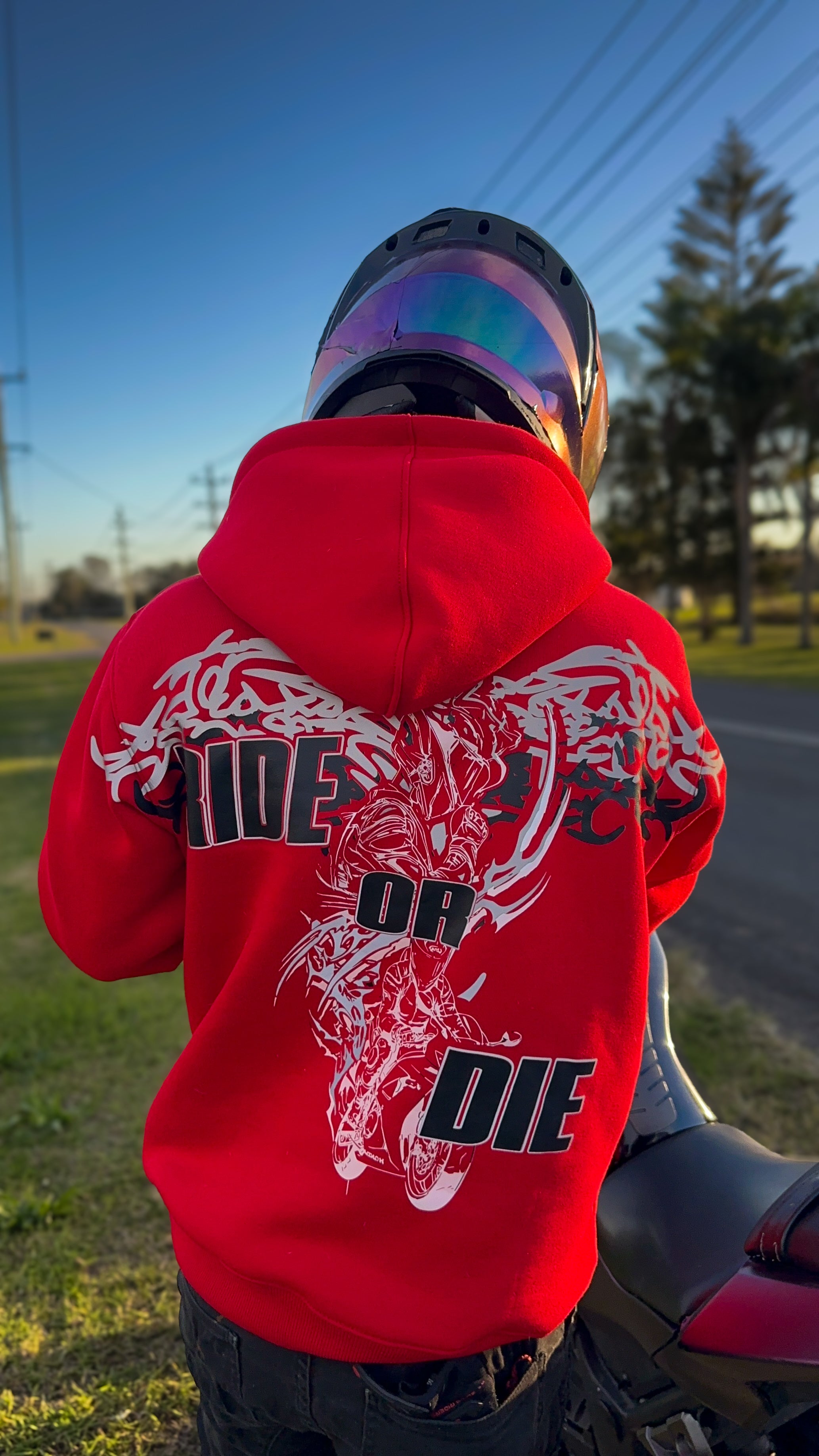 A motorbike rider viewed from behind wearing a bright red hoodie with a large white and black graphic on the back, standing outdoors near a motorcycle on a road with trees and a blue sky in the background.