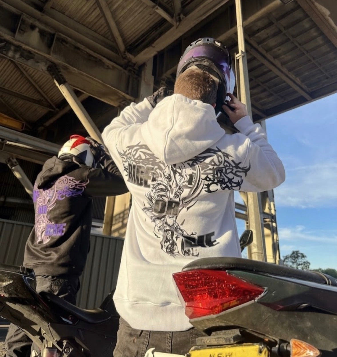 A person wearing a white hoodie with a large black graphic on the back stands under a metal structure near a motorcycle, holding a phone to their face. Another person in a black hoodie with purple graphics is visible in the background.
