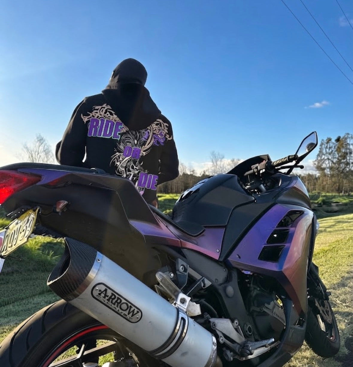 A person wearing a black hoodie with a purple graphic stands next to a purple and black motorcycle outdoors under a clear blue sky. The motorcycle has a visible silver ARROW exhaust system.